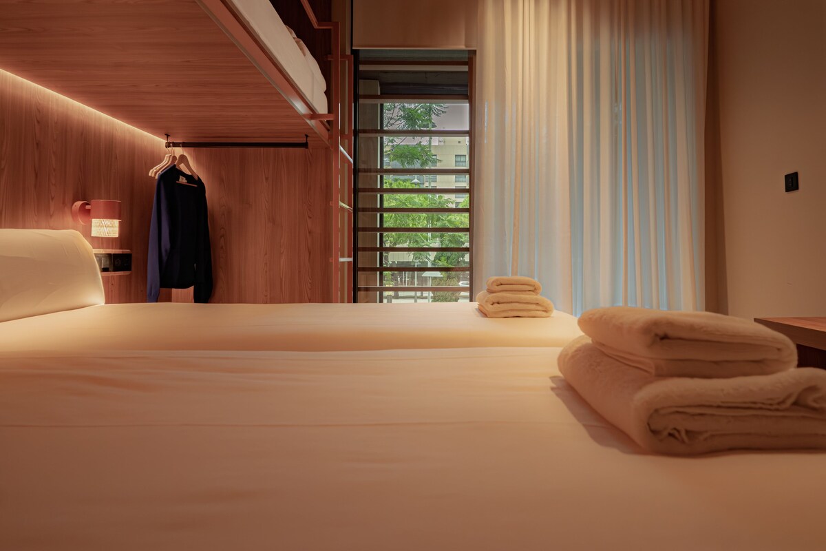 A hotel room with a bed, a hanging coat rack, a window with curtains, and a view of greenery outside.