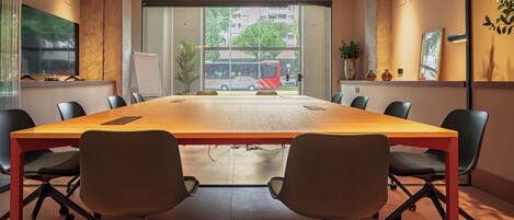 A conference room with a long table, chairs, a whiteboard, and large windows with a view of buildings and trees.