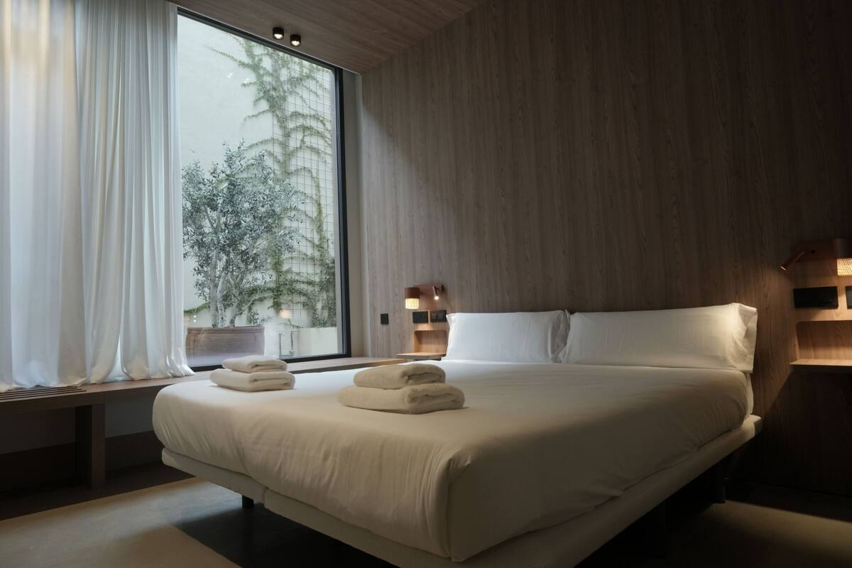 A modern hotel room with a large bed, a window with curtains, a bedside table with a lamp, and a view of trees outside.