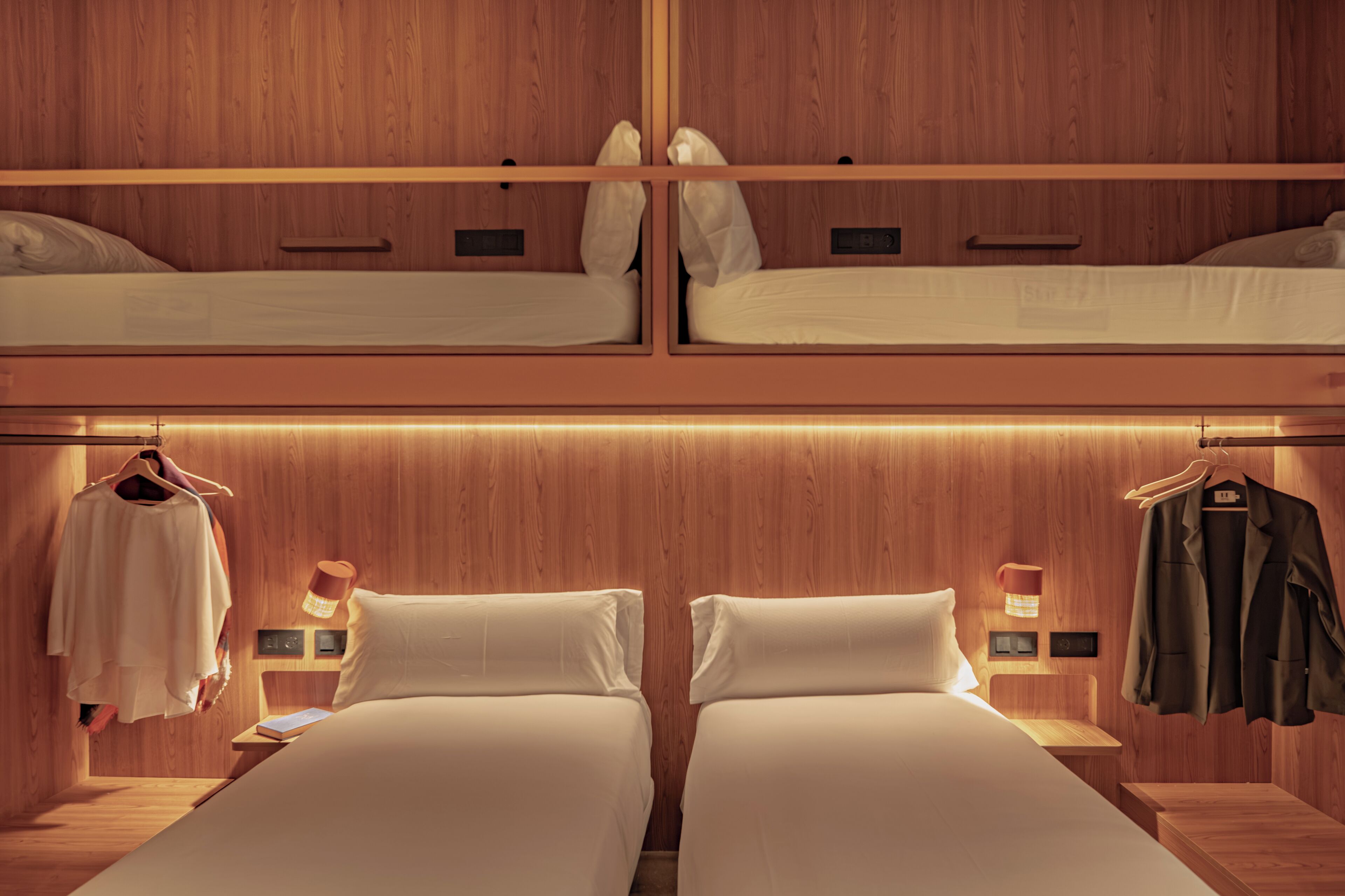 Habitación con literas, paredes de madera, dos mesitas de noche y ropa colgada.