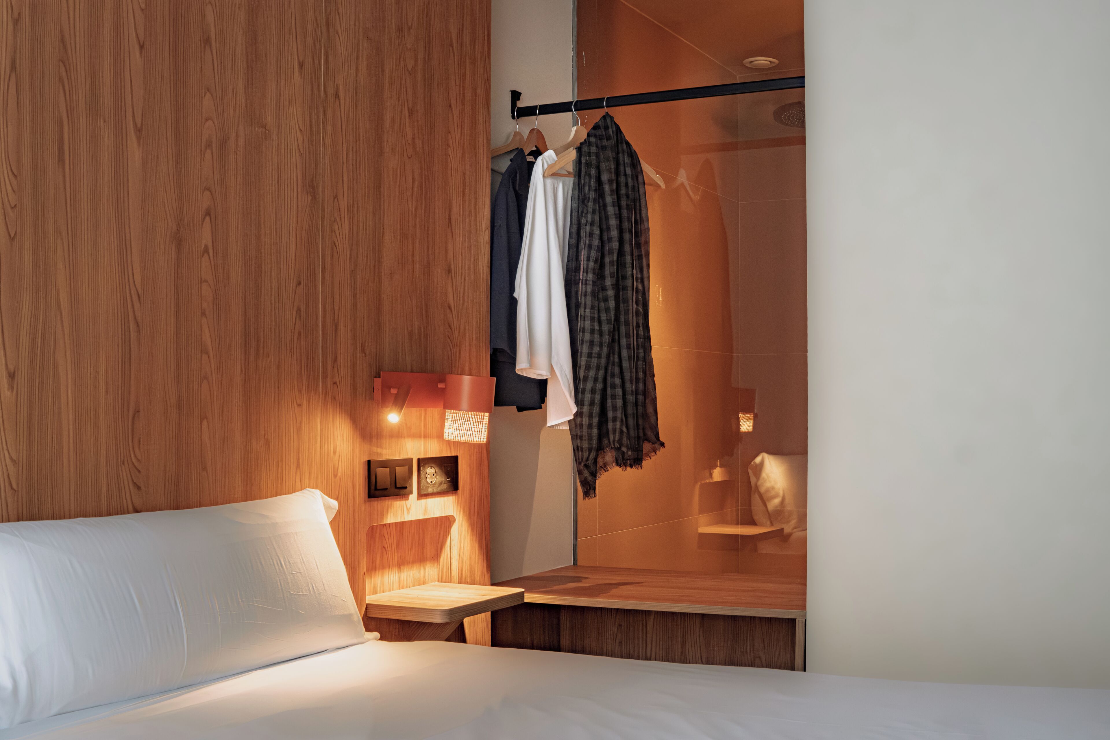 Habitación de hotel con pared de madera, cama, mesita de noche, espejo y perchero con varias prendas.