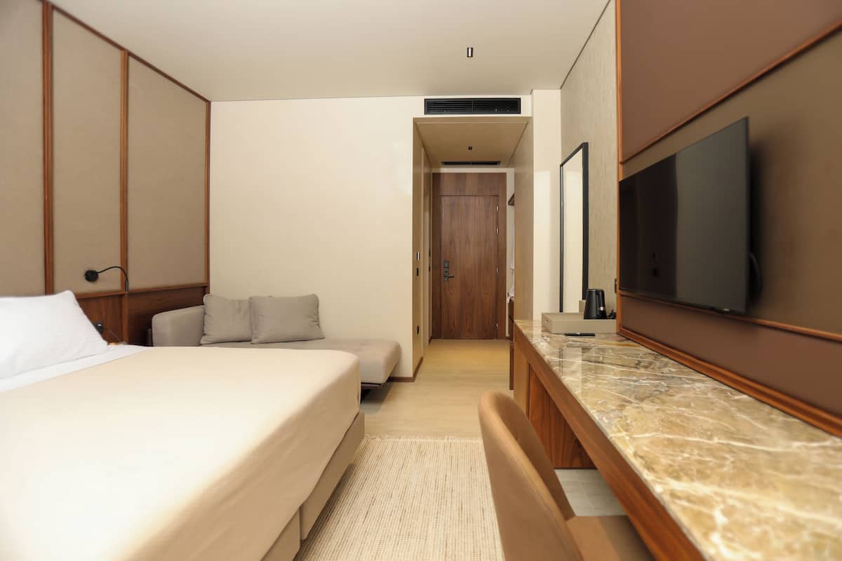 Una habitación de hotel moderna con una cama grande, un televisor de pantalla plana montado en la pared, un armario de madera y una pequeña zona de estar con cojines.
