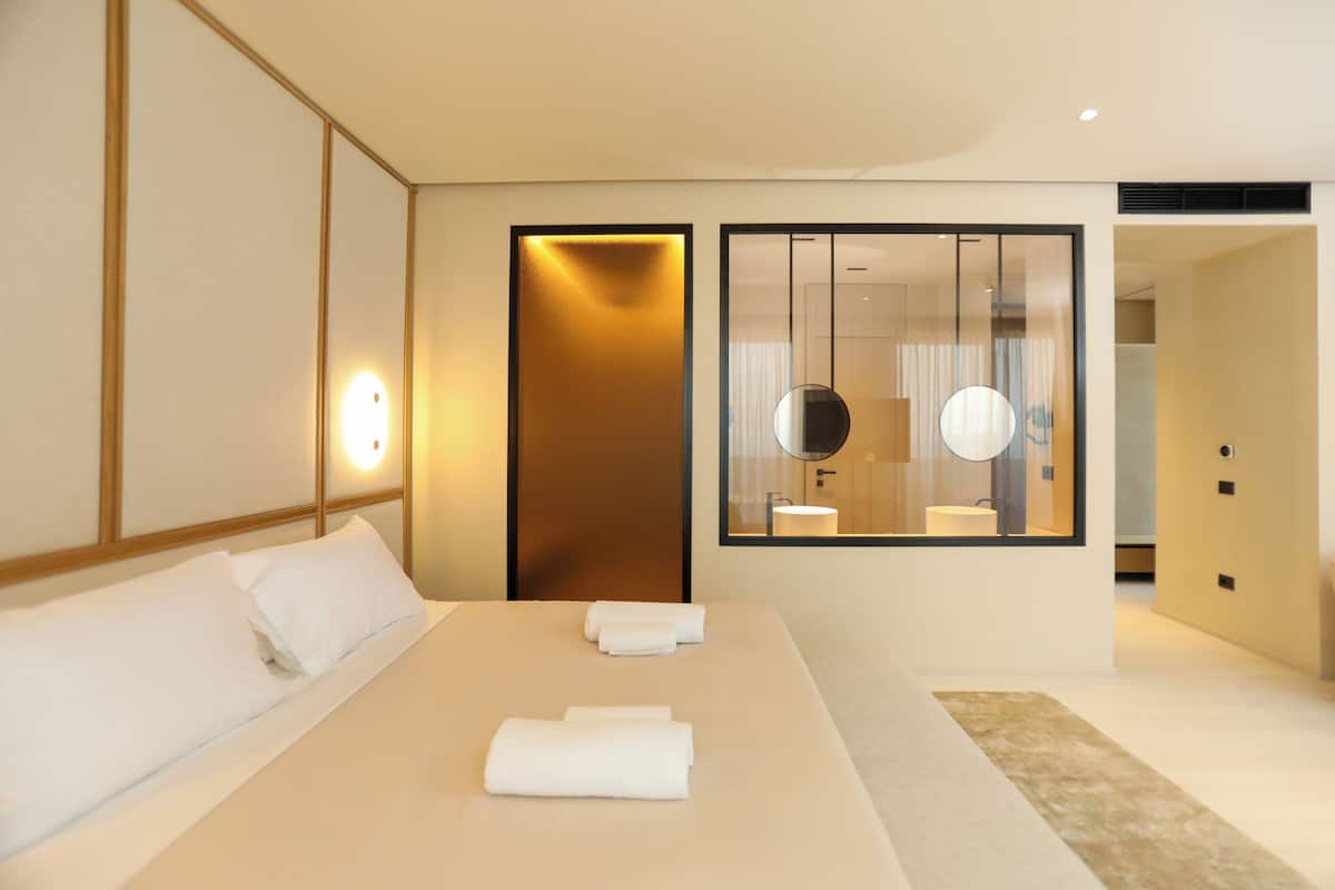 Habitación de hotel moderna con una cama grande, una mesita de noche, un espejo y vistas al baño a través de un separador de vidrio.