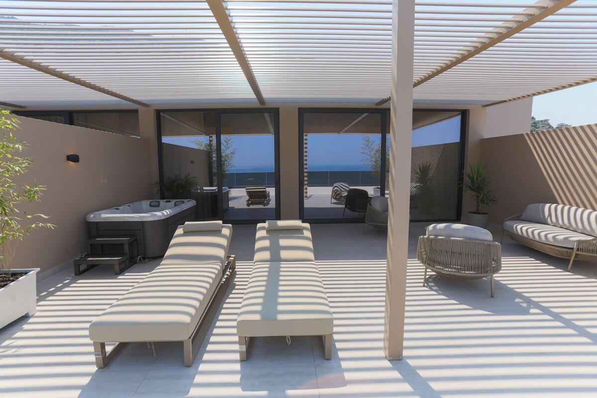 A modern outdoor lounge area with sunbeds, a hot tub, and a view of the sea.