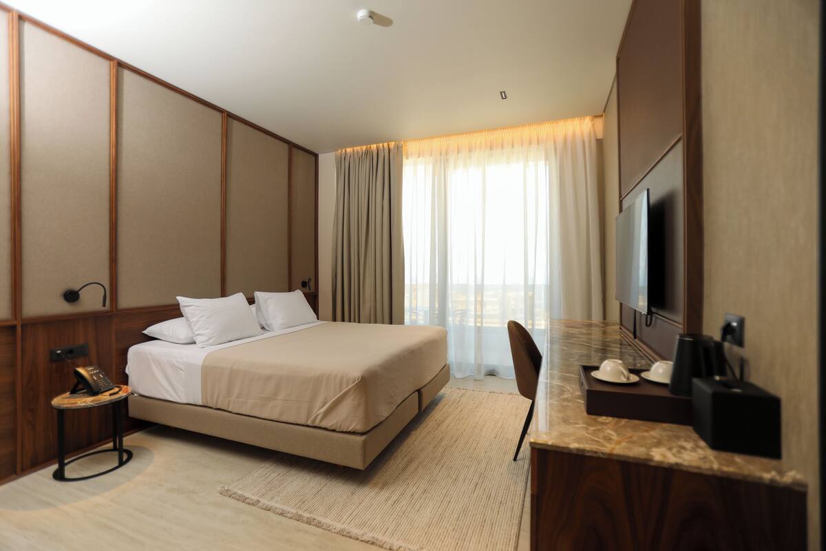Una habitación de hotel con una cama grande, un escritorio con silla, televisor y ventana con cortinas.