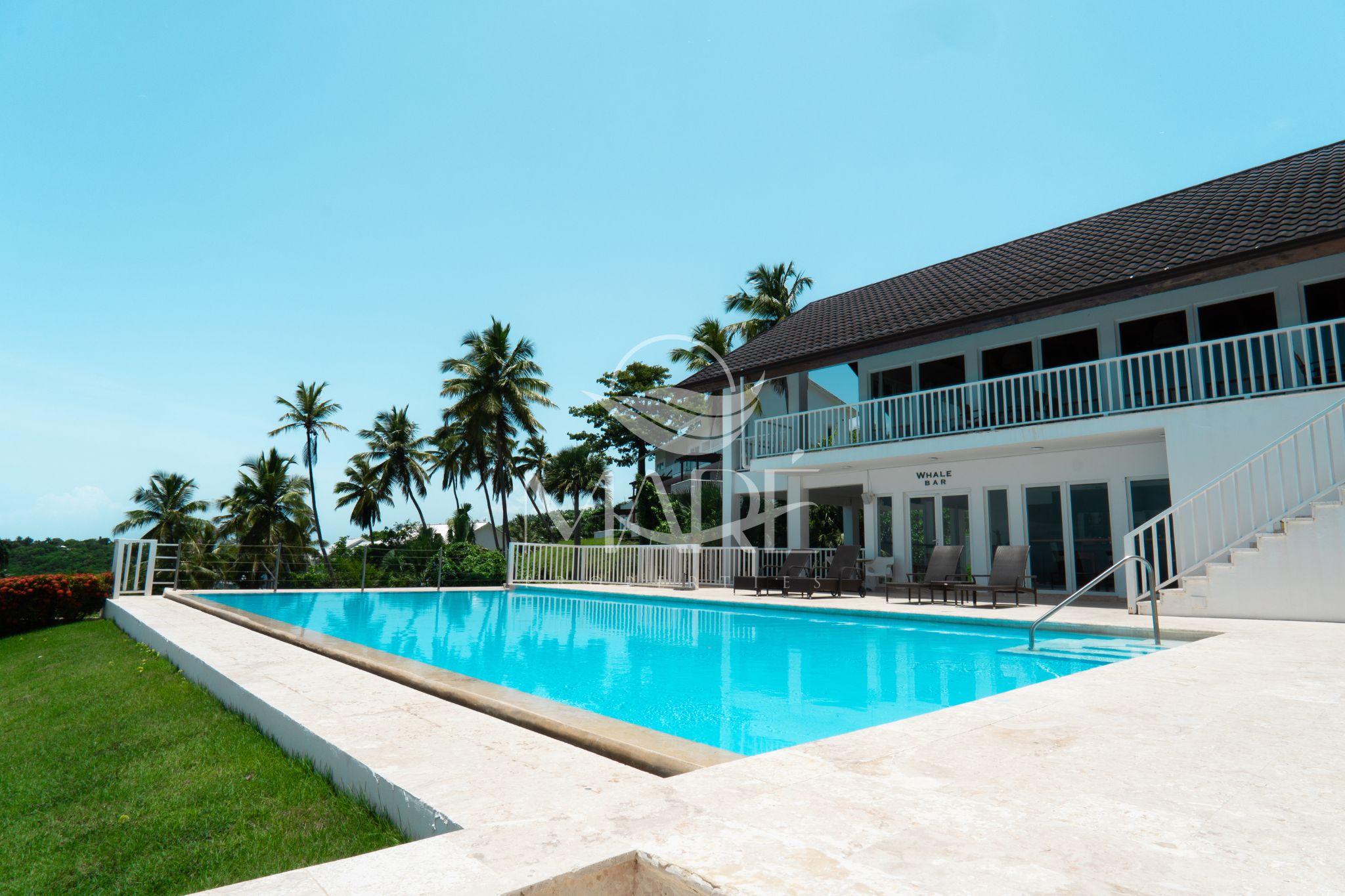 Ein großer Pool mit weißer Liegefläche, eine Bar namens 'Whale Bar' und ein zweistöckiges Gebäude mit Balkon.