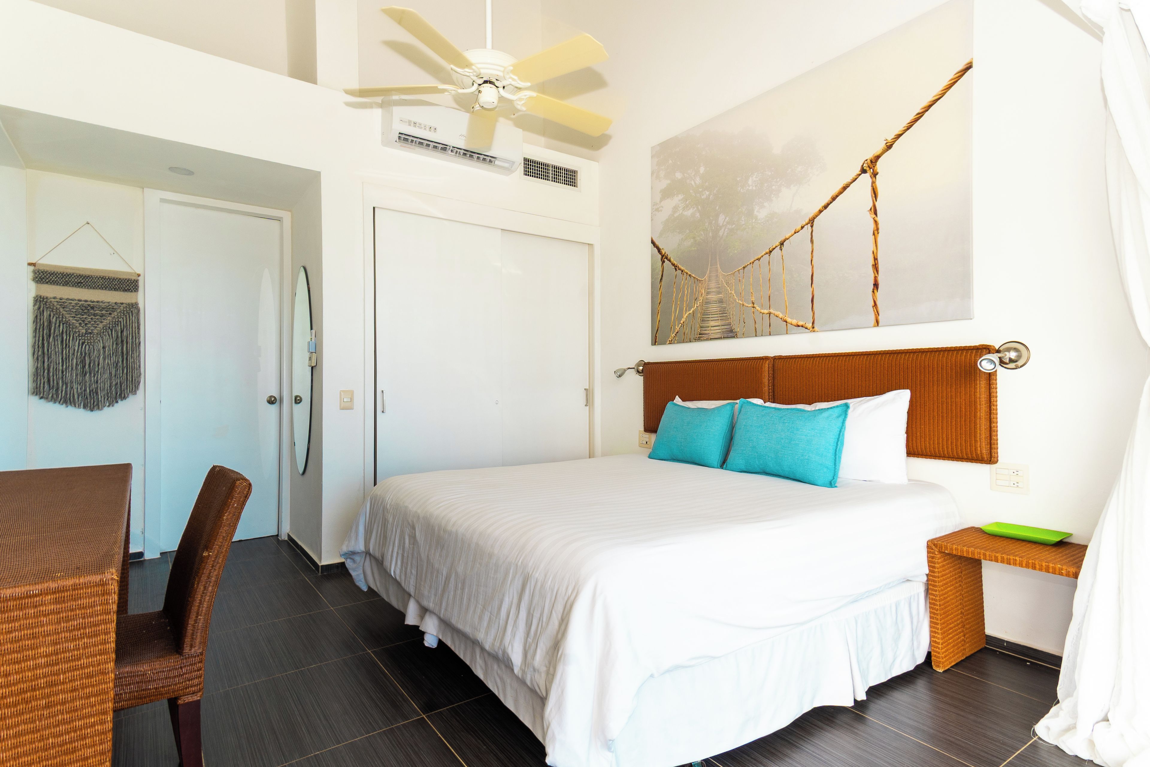 Une chambre d’hôtel comprenant un lit, une chaise, une table, un ventilateur de plafond, un climatiseur fixé au mur et un tableau représentant un pont.