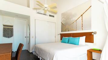 A hotel room with a bed, a chair, a table, a ceiling fan, a wall-mounted air conditioner, and a painting of a bridge.