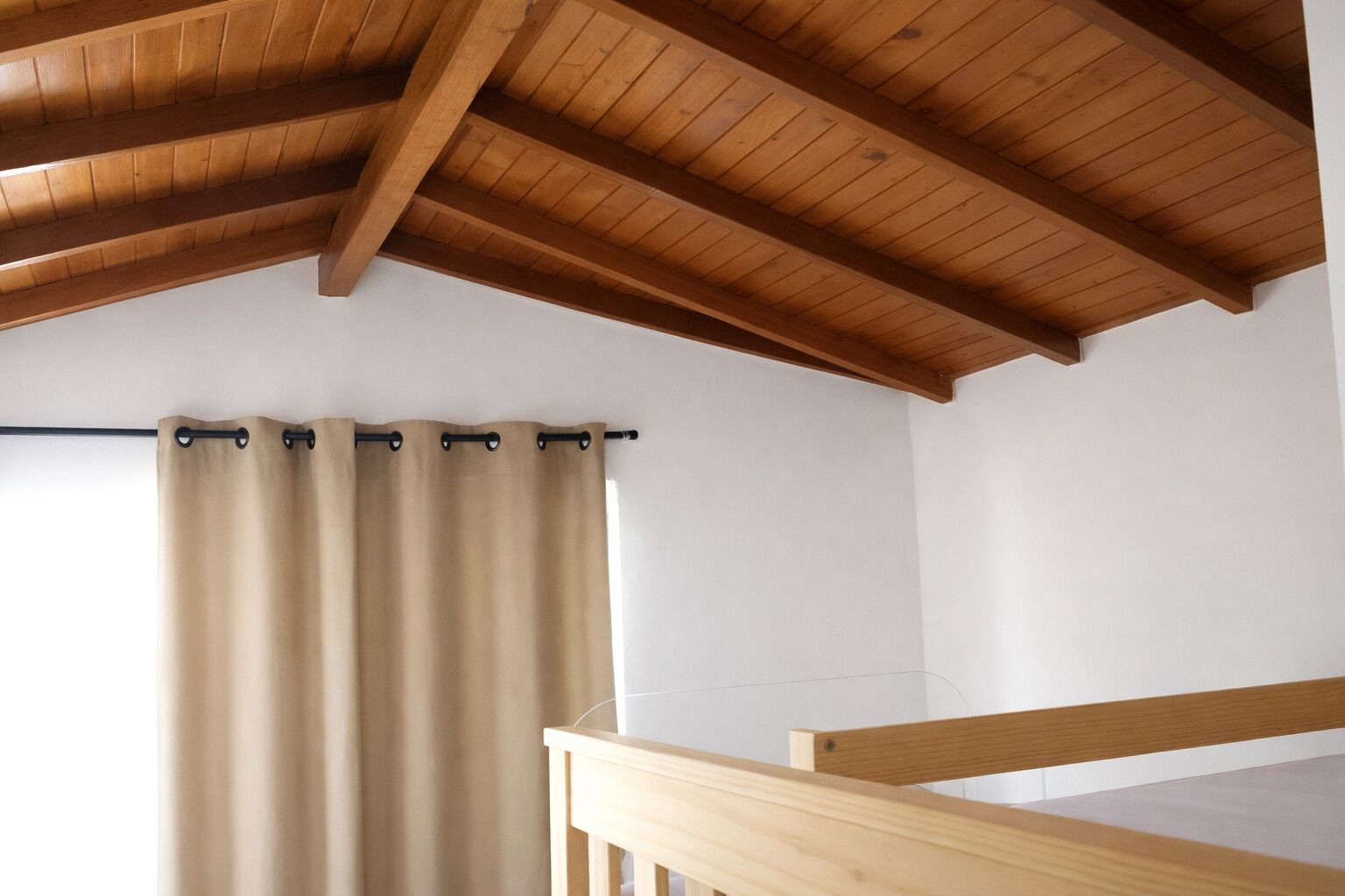 Una habitación con techo de madera, cortina color beige y baranda de madera.