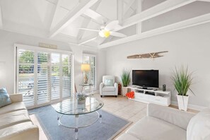 A modern living room with a glass coffee table, a white sofa, a TV on a white stand, and a ceiling fan.