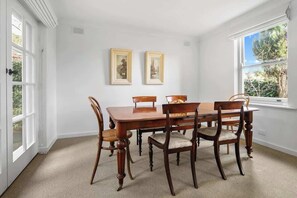 Comedor con mesa y sillas de madera, dos cuadros en la pared y ventana con vista a follaje.