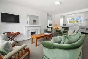 A living room with a fireplace, green upholstered furniture, a flat-screen TV, and a kitchen area with a window. - Between City & Hills (Mitcham)