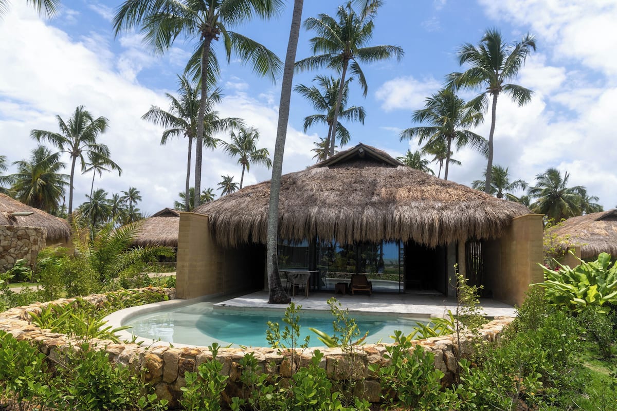 Una zona de piscina con construcciones de techo de paja, rodeada de palmeras y vegetación.