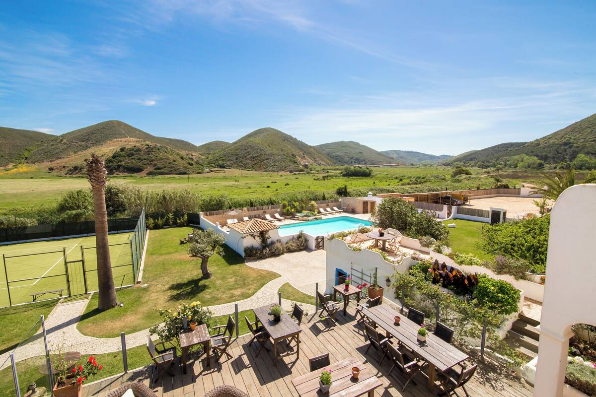 A pool area with a view of hills, outdoor seating, and a tennis court.