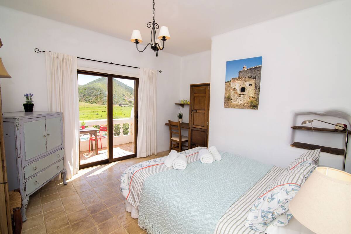 A bedroom with a bed, a desk, a chair, a dresser, a window with curtains, and a view of a green landscape.