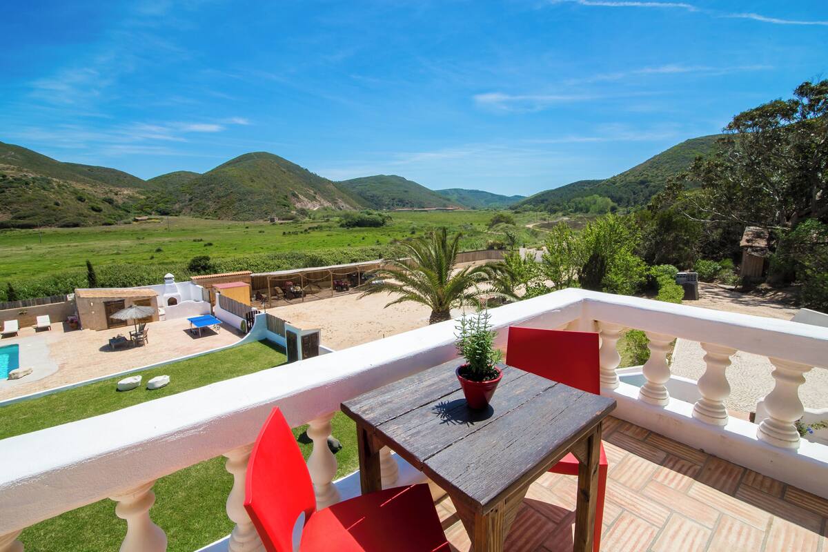 A balcony with a wooden table and red chairs, overlooking a pool and a lush green landscape with hills.