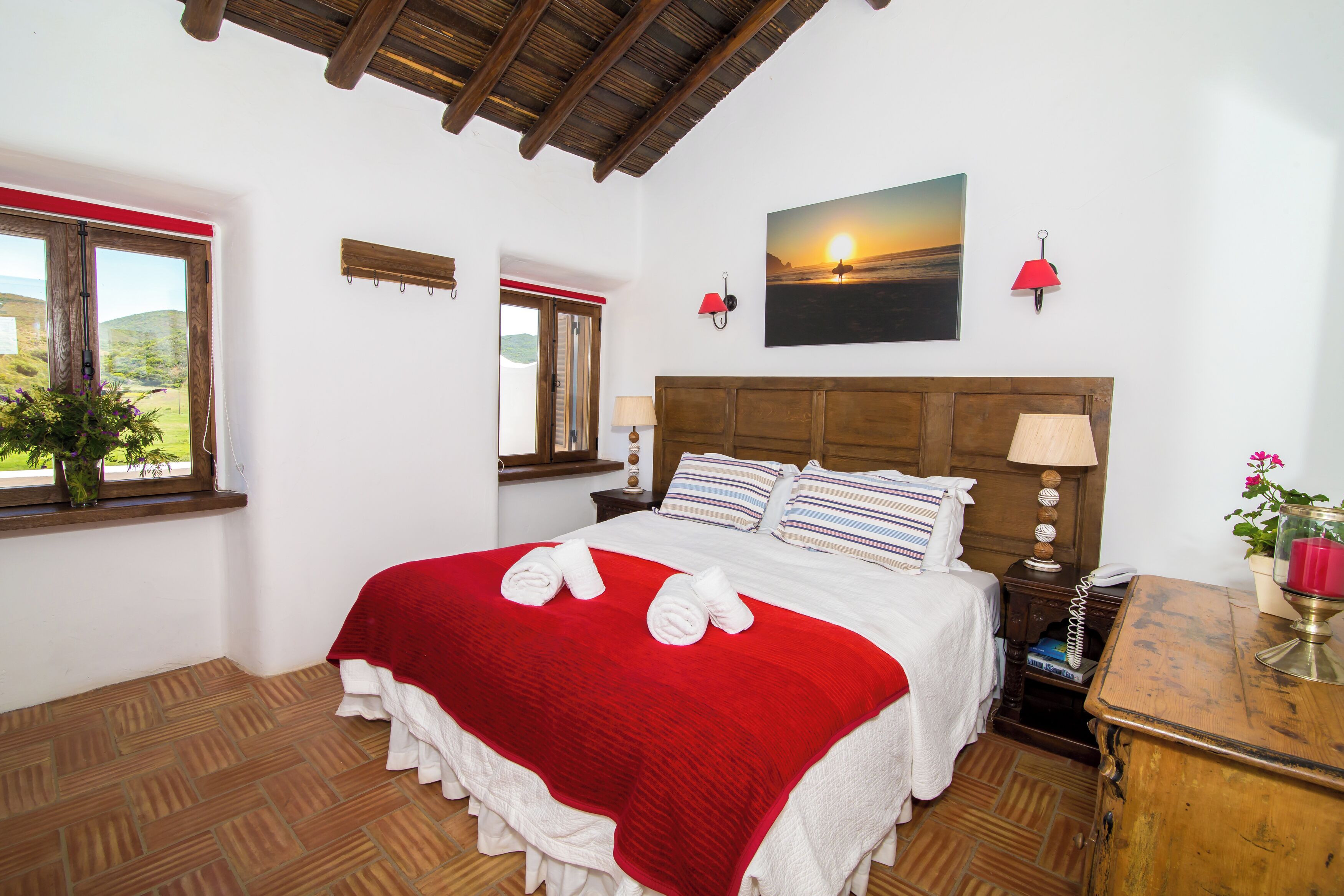 A neatly made bed with a red and white bedspread, two pillows, and two rolled towels. There is a wooden headboard, a nightstand with a lamp, and a painting of a sunset on the wall.
