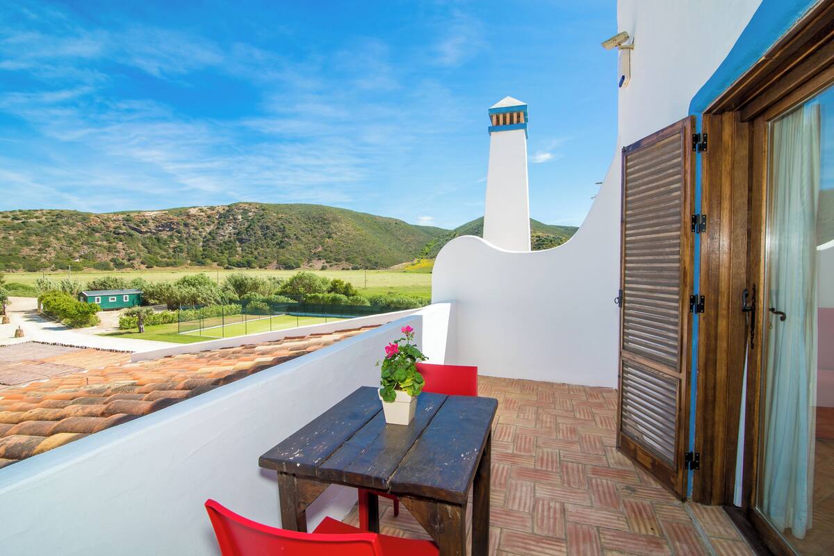 A balcony with a table and chairs, a view of the countryside, and a building with a tower.