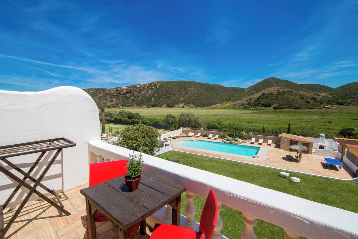 A balcony with a view of a swimming pool, a table, and red chairs.