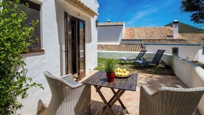 Eine Terrasse mit Korbmöbeln, einem Tisch mit Topfpflanze und Obst, und Blick auf ein Gebäude mit Ziegeldach.