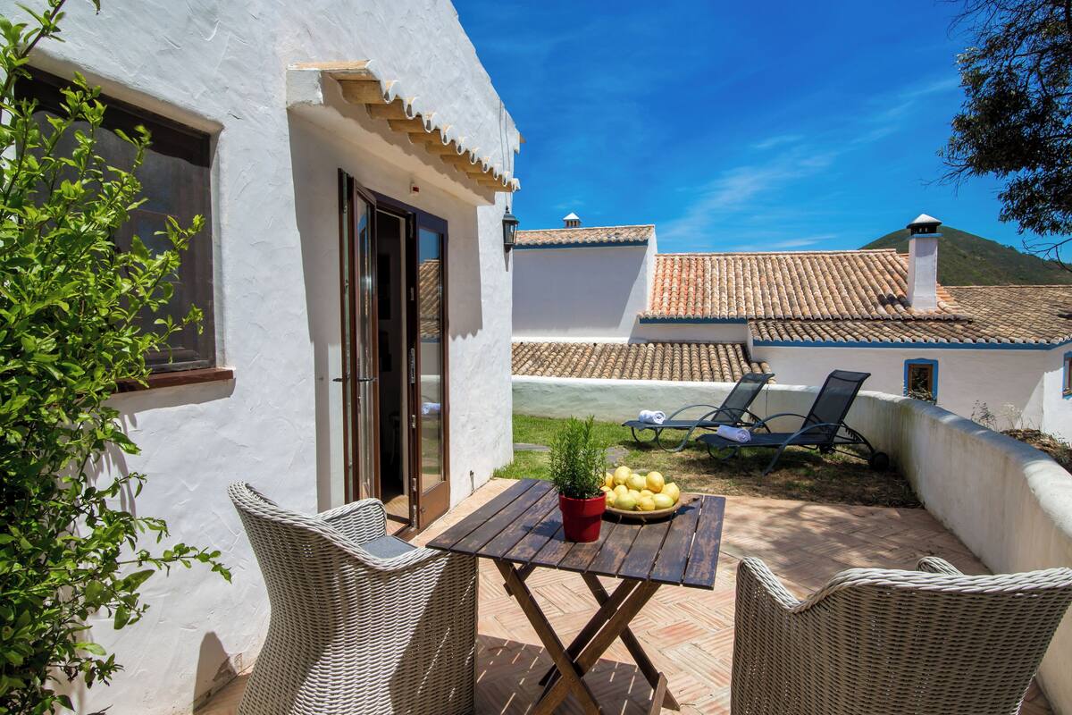 A patio with wicker furniture, a table with a potted plant and fruit, and a view of a building with a tiled roof.