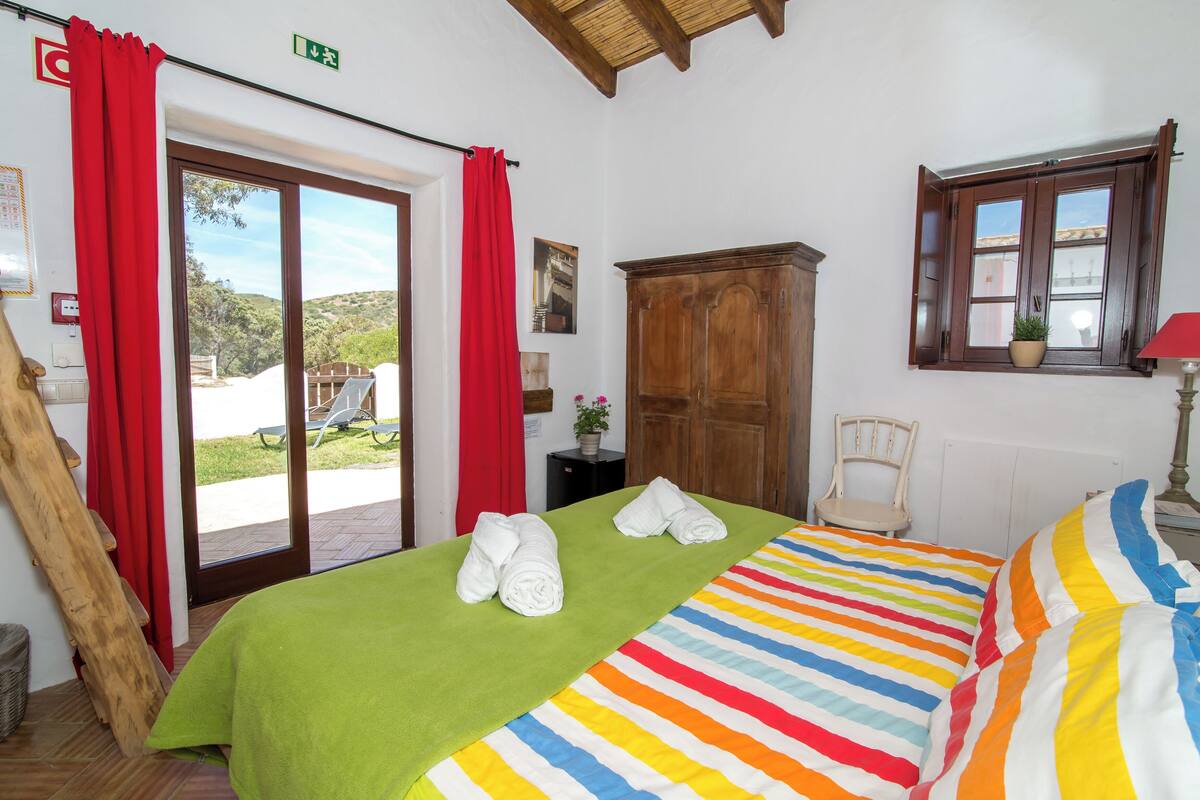 A hotel room with a green bedspread, striped pillows, a wooden wardrobe, a window with a view, and a red curtain.