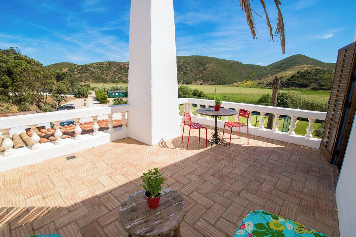 A balcony with a view of a green landscape, a table with chairs, and a small potted plant.