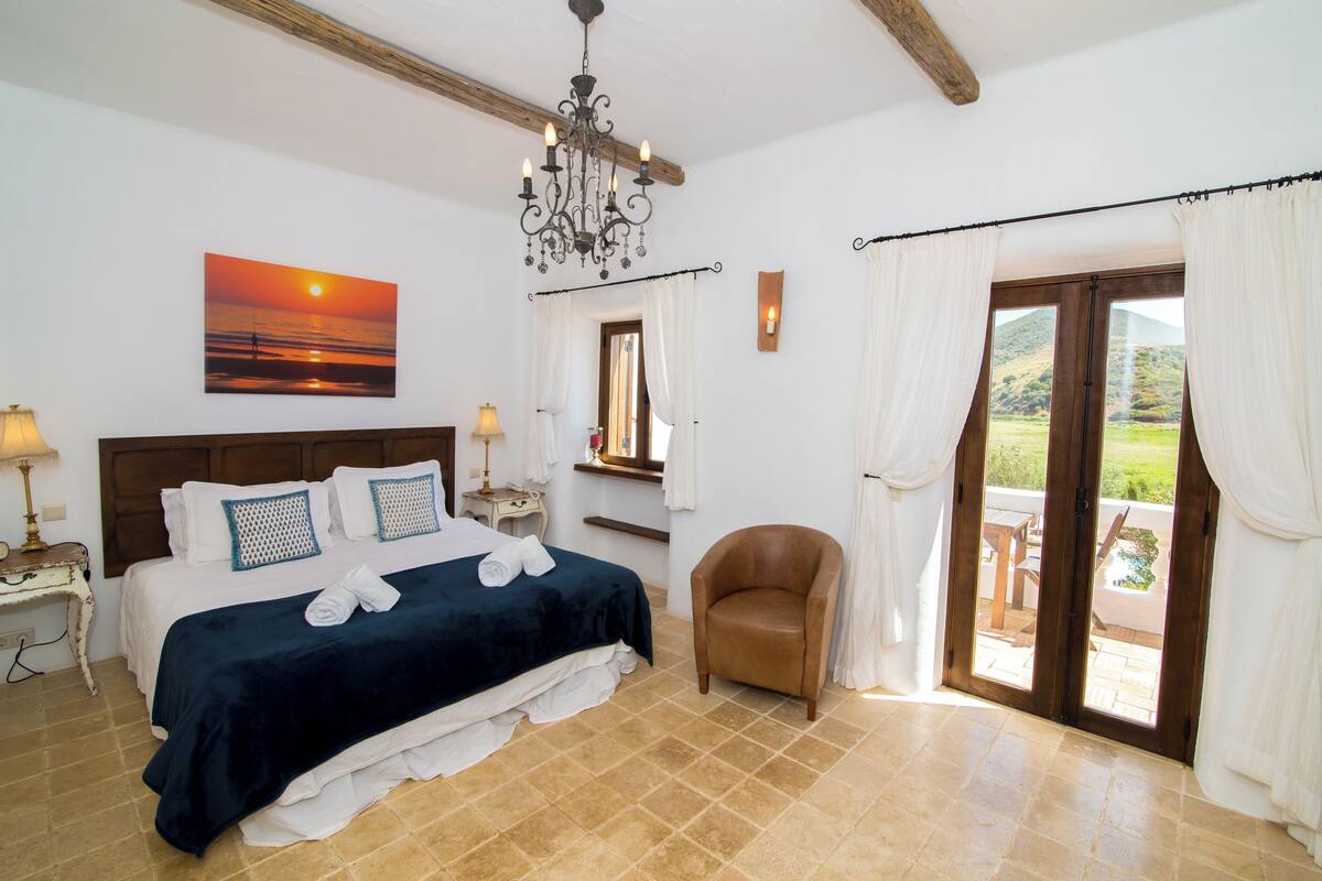 A bedroom with a bed, a chair, a chandelier, a painting, and a view of the outdoors.