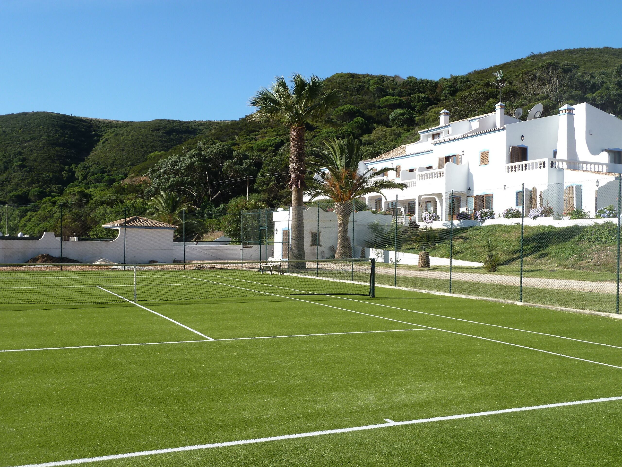 A tennis court with a net, surrounded by a fence. In the background, there is a white building with balconies and satellite dishes.