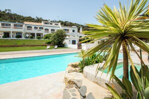 A swimming pool with a stone border, surrounded by a well-maintained lawn and a large white building with multiple balconies in the background. - Casa Fajara (Aljezur)