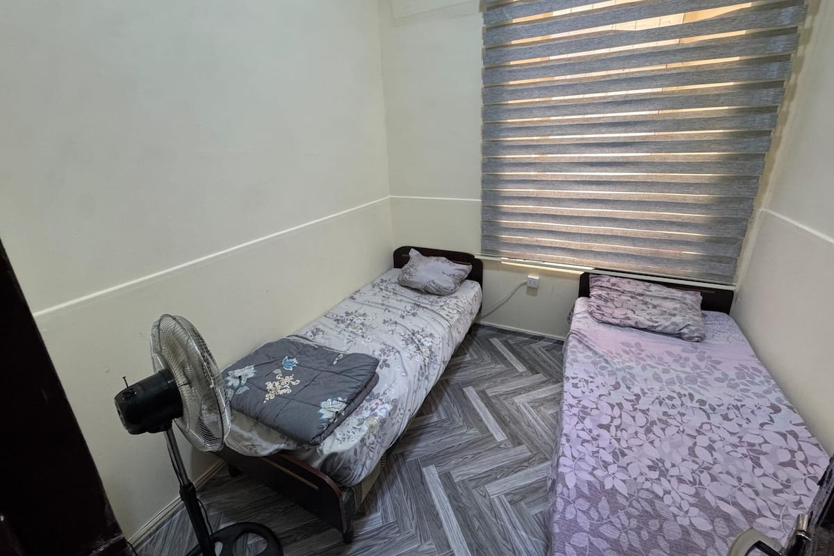 A small, compact room with two single beds, a standing fan, and a window with blinds.