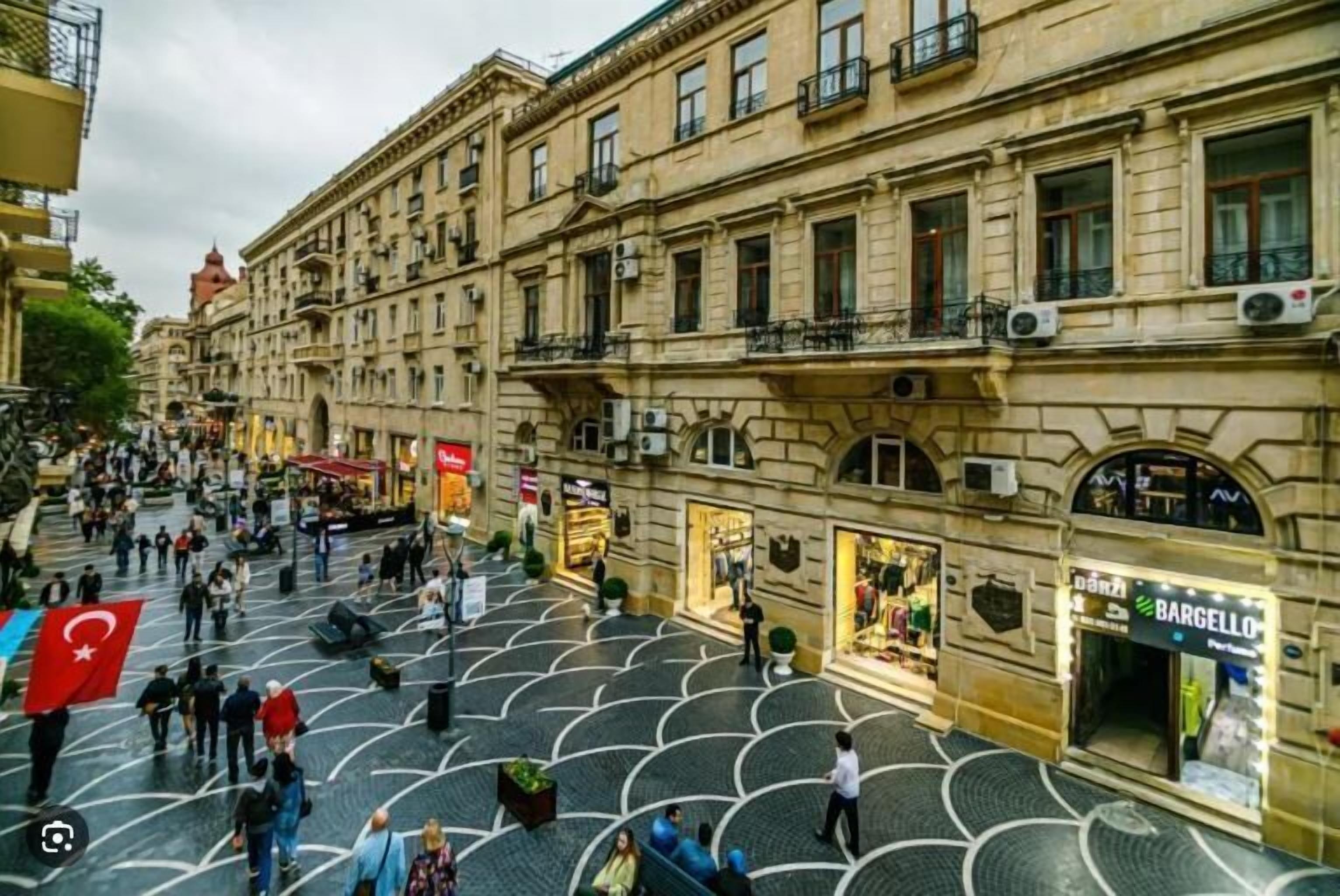 Une vaste place publique, dotée d’un sol carrelé, entourée de bâtiments historiques pourvus de balcons et de devantures de magasins.