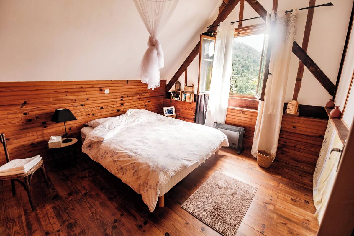 A bedroom with a wooden floor, a bed with a white bedspread, a nightstand with a lamp, a window with curtains, and a view of the outdoors.