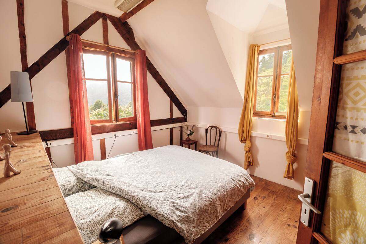 A bedroom with a sloped ceiling, wooden beams, a bed with a white blanket, a small table with a plant, and a window with curtains.