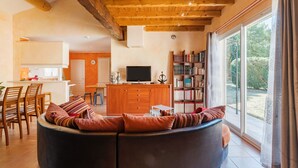A modern living room with a curved sofa, wooden ceiling beams, a television, a bookshelf, and a large window. - Villa 'Le Verger Du Midi' with pool, private terrace, and Wi-Fi (Cadenet)