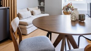 Ein modernes Wohnzimmer mit einem runden Holzesstisch, zwei grauen Polsterstühlen, einem beigen Sofa mit Kissen, einer Holzvertikalvorrichtung, einem Bücherregal und einer Pendelleuchte.