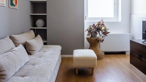 Ein modernes Wohnzimmer mit grauem Sofa, einem kleinen Hocker, einem Bücherregal und einem Fenster mit Tageslicht.