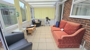 A sunroom with a sofa, a table, a telescope, and a window overlooking a garden. - Herne Bay - House - Sleeps 7 - Parking (Herne Bay)