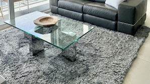 A modern living room with a glass coffee table, a black leather sofa, and a view of the cityscape through large windows.