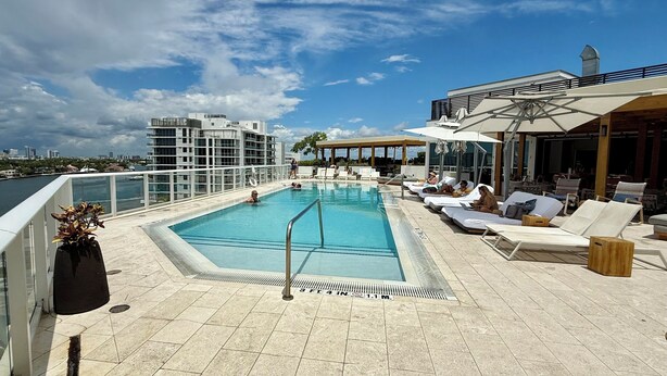 A rooftop pool with lounge chairs and umbrellas, surrounded by modern buildings and a clear sky.