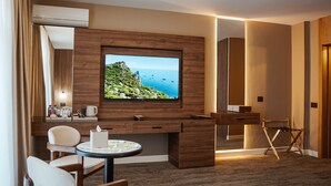 Habitación de hotel con un televisor de pantalla plana grande montado en una pared de madera, un escritorio con una silla, una pequeña mesa redonda con una vela y una silla plegable.