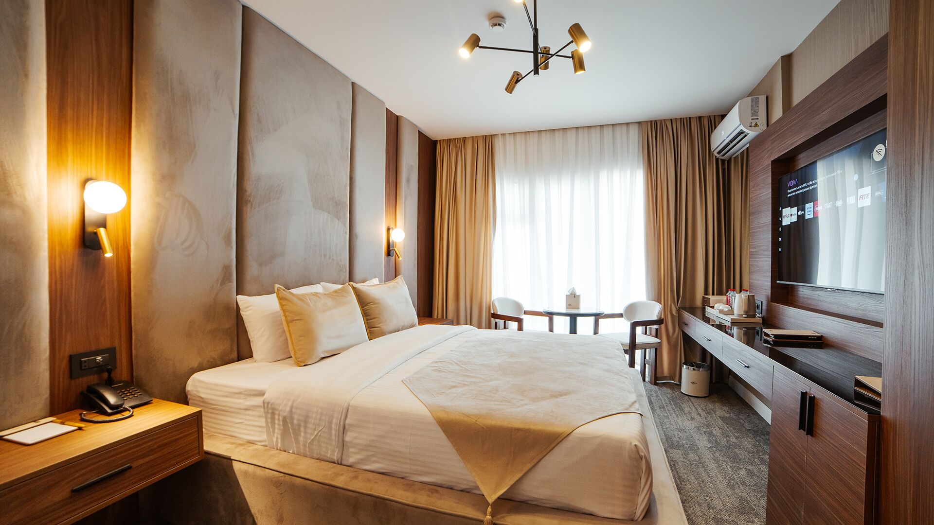 Une chambre d’hôtel avec un grand lit, une table de chevet avec un téléphone, un bureau avec une chaise, une télévision et une fenêtre avec des rideaux.