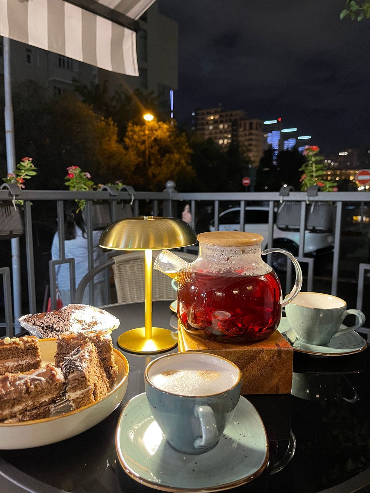 A table set with a teapot, a cup of coffee, and a slice of cake on a balcony with a city view at night.