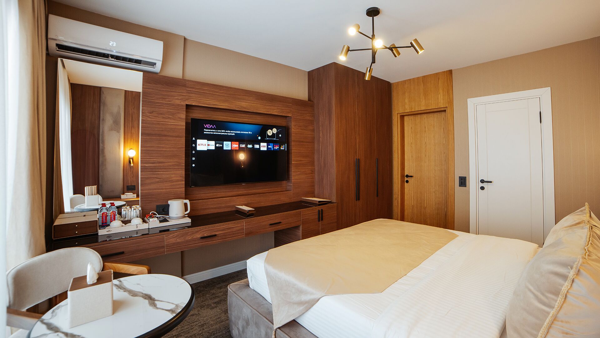 A hotel room with a bed, a desk with a TV, a chair, a coffee maker, and an air conditioning unit.