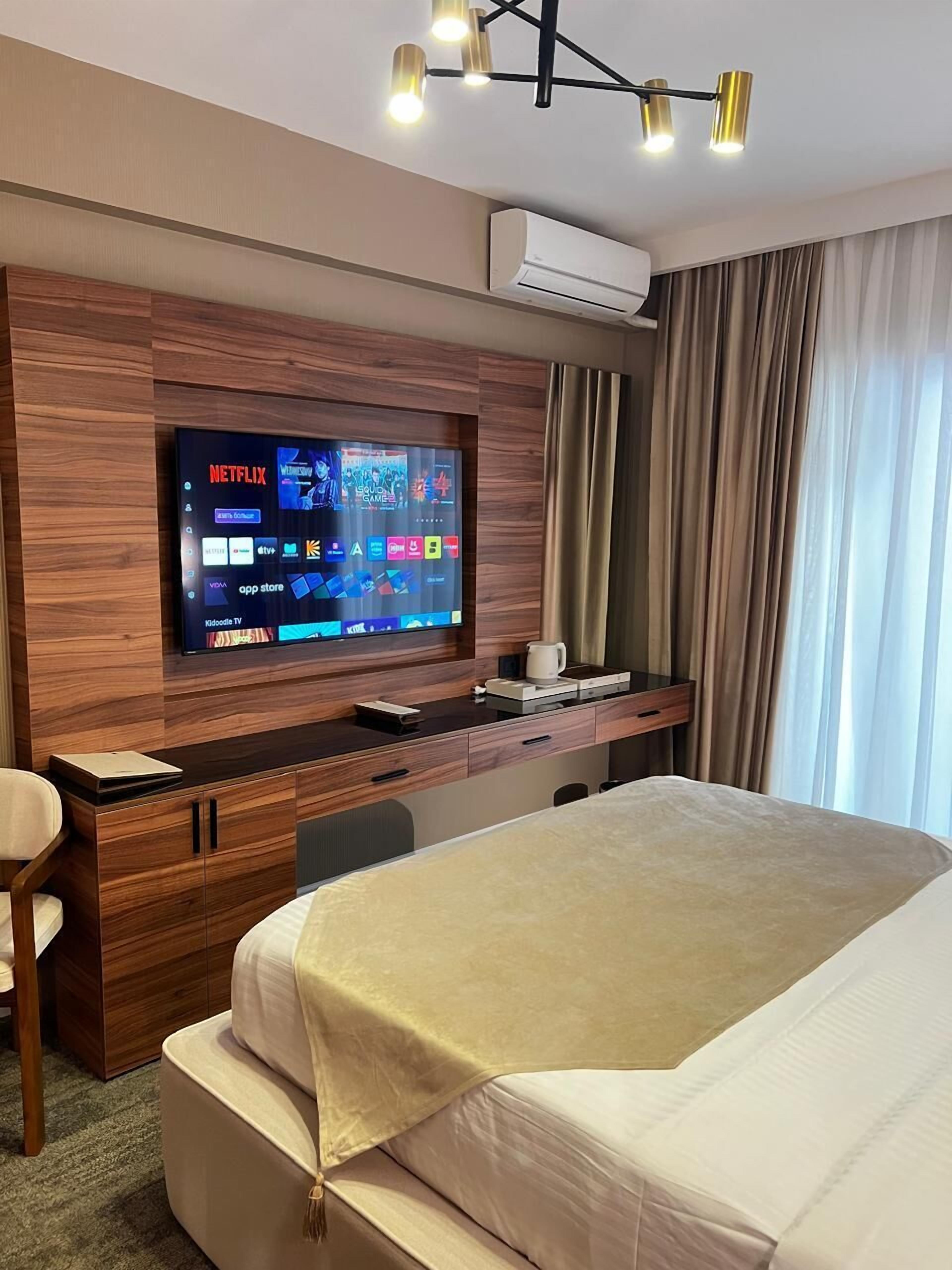 Habitación de hotel con una televisión de pantalla plana grande montada en una pared de madera, una cama con cubierta color beige, un escritorio con cafetera y una ventana con cortinas.