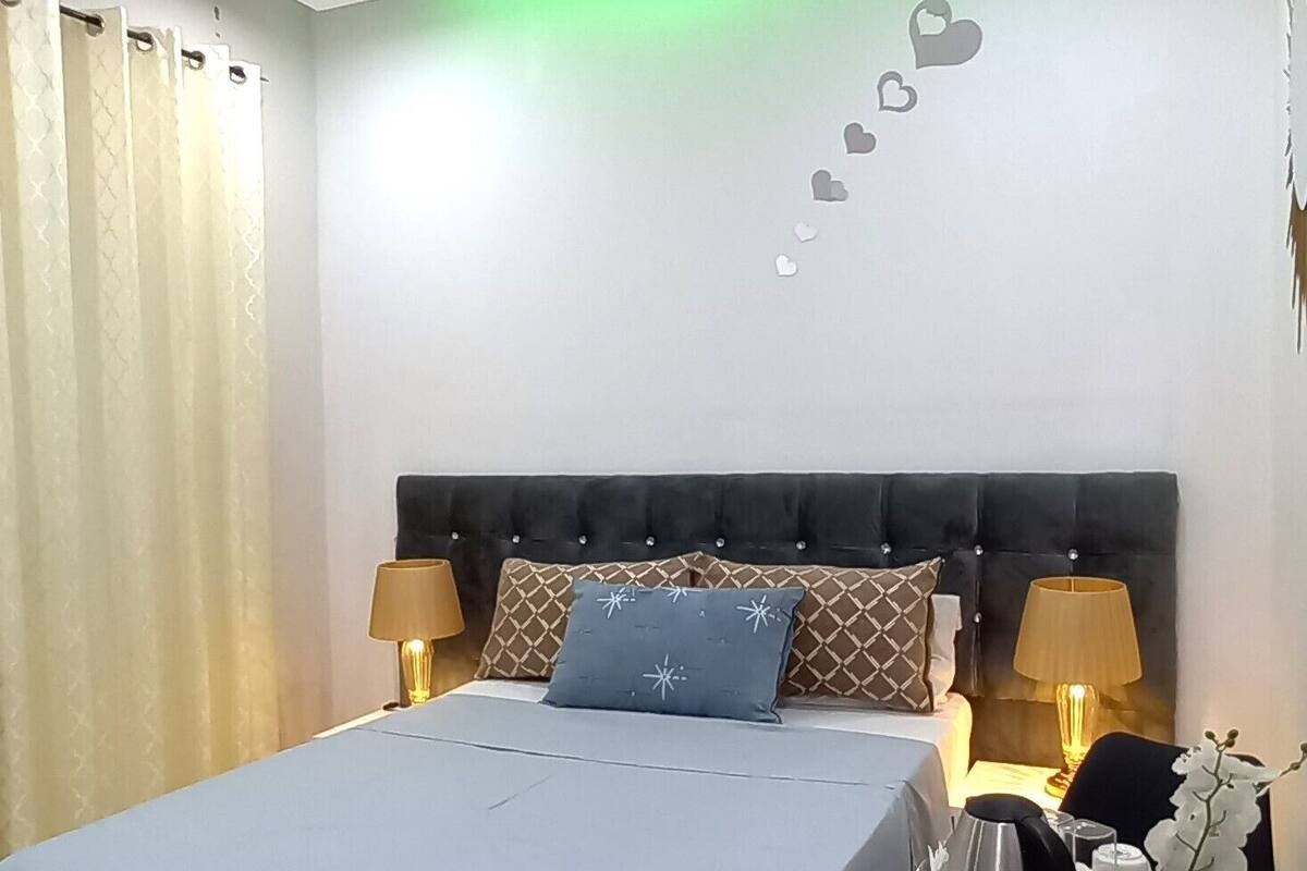 Ein Hotelzimmer mit einem Bett, zwei Nachttischen mit Lampen, einem kleinen runden Tisch mit Wasserkocher und Tablett sowie einer Wand mit herzförmiger Dekoration.
