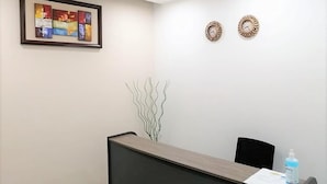 A reception area with a wooden reception desk, a black chair, a framed abstract painting, two decorative wall clocks, and a decorative plant. - 4040 Accomodation (Matola)