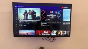 A flat-screen TV mounted on a wall displaying a YouTube interface with various video thumbnails and a remote control on a stand below. - 4040 Accomodation (Matola)