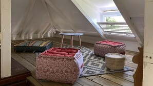 Ein gemütliches Dachzimmer mit schrägen Wänden, zwei gemusterte Hocker, ein kleiner runder Tisch und ein Fenster mit Blick auf Grünflächen.