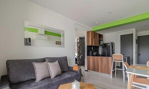 A modern living room with a grey sofa, a coffee table, and a dining area with a glass table and chairs. A kitchen is visible in the background with wooden cabinets and a microwave. - Epic Suite 513 Deluxe Aeropuerto (Pereira)